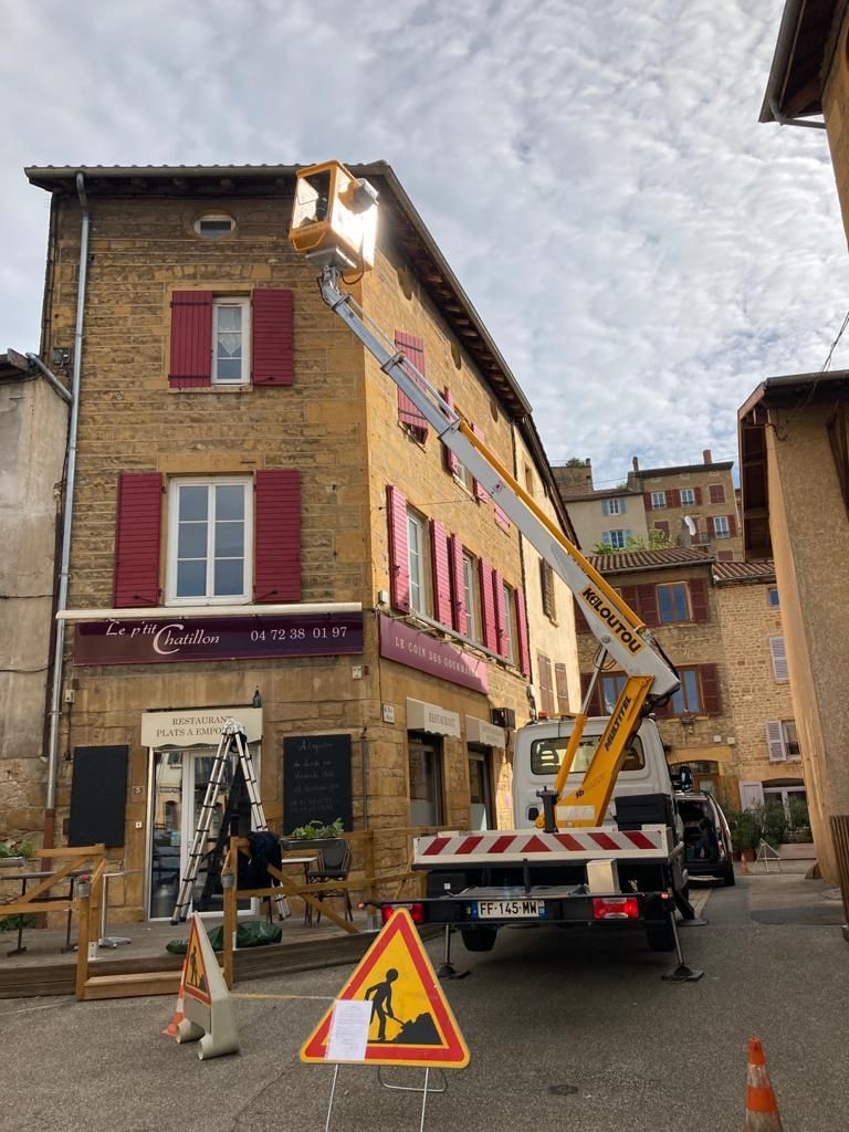 Un camion utilitaire jaune équipé d'une nacelle élévatrice est stationné sur une place pavée, devant un bâtiment historique en pierre.