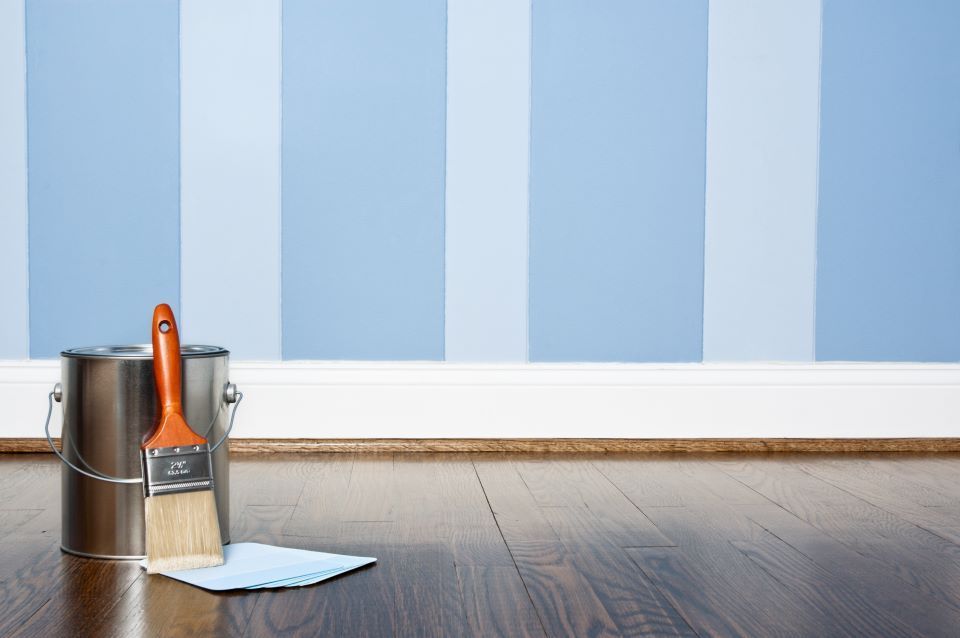 Ein Eimer Farbe und ein Pinsel stehen auf dem Boden vor einer blau-weiß gestreiften Wand.