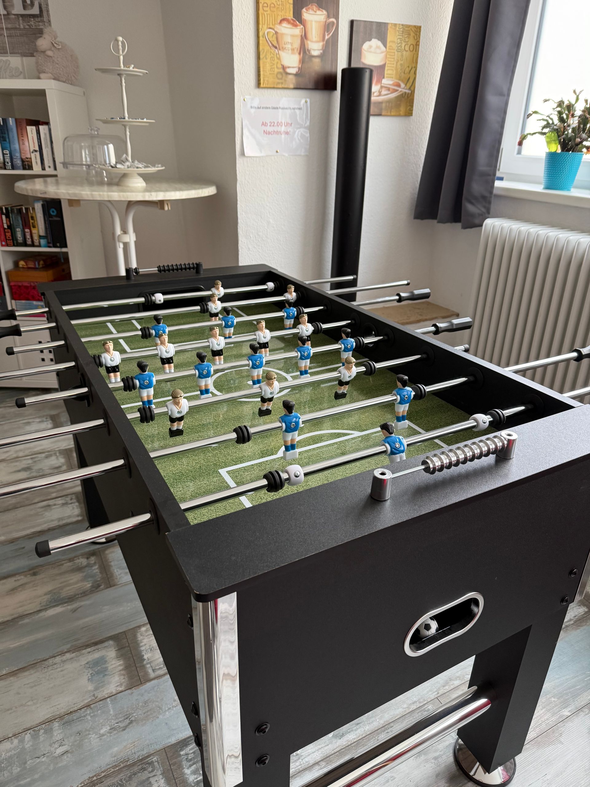 Tischkicker in einem hellen Raum mit Stühlen, Vorhängen, Heizkörper und Wandbildern im Hintergrund