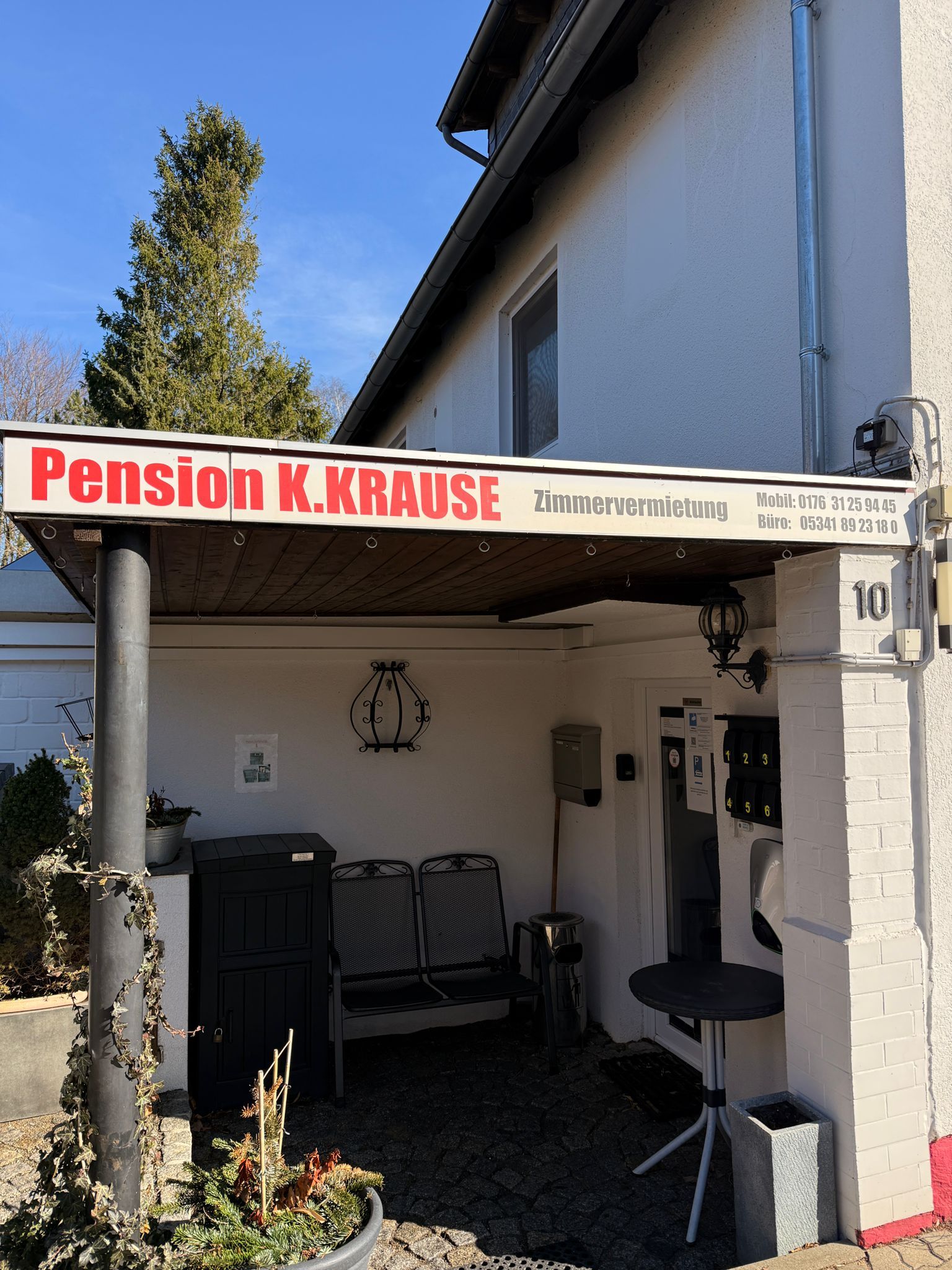 Eingang zur Pension K.KRAUSE mit weißer Markise, rotem Schild und kleinem Sitzbereich im Freien