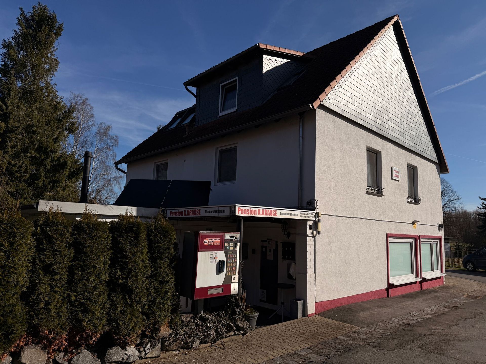 Zweistöckiges Haus mit schrägem Dach, hellen Wänden, rot umrandeten Fenstern und Hecken an der Vorderseite.