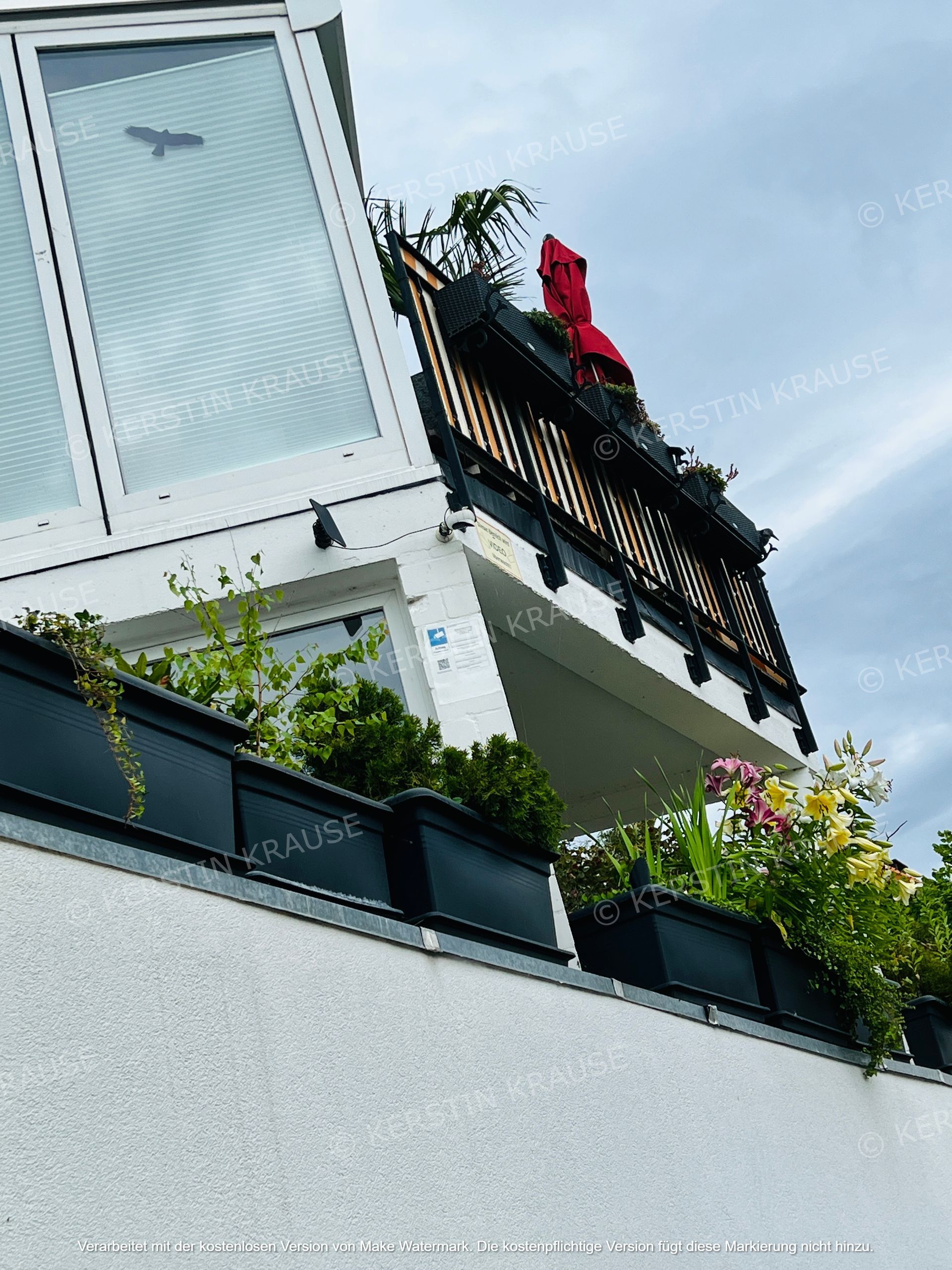 Balkon mit schwarzem Geländer, Topfpflanzen und einem roten Handtuch an einem weißen Gebäude vor blauem Himmel