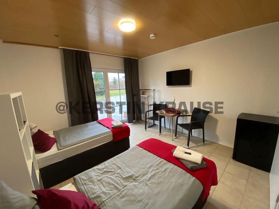 Kleines Schlafzimmer mit zwei Einzelbetten, Schreibtisch, Wandfernseher und Balkontüren, dekoriert in Grau, Weiß und Rot.