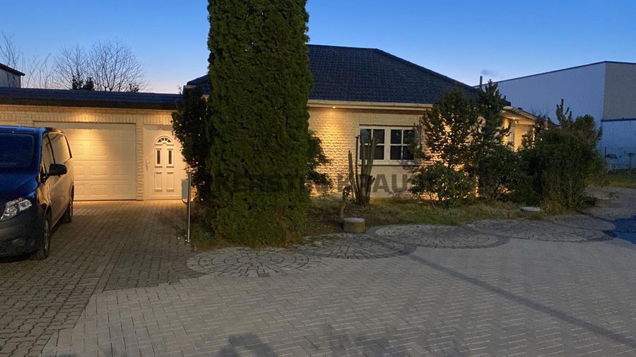 Ein eingeschossiges Haus in der Abenddämmerung mit beleuchteten Fenstern, einem hohen Baum davor und einem geparkten Auto in der Einfahrt.