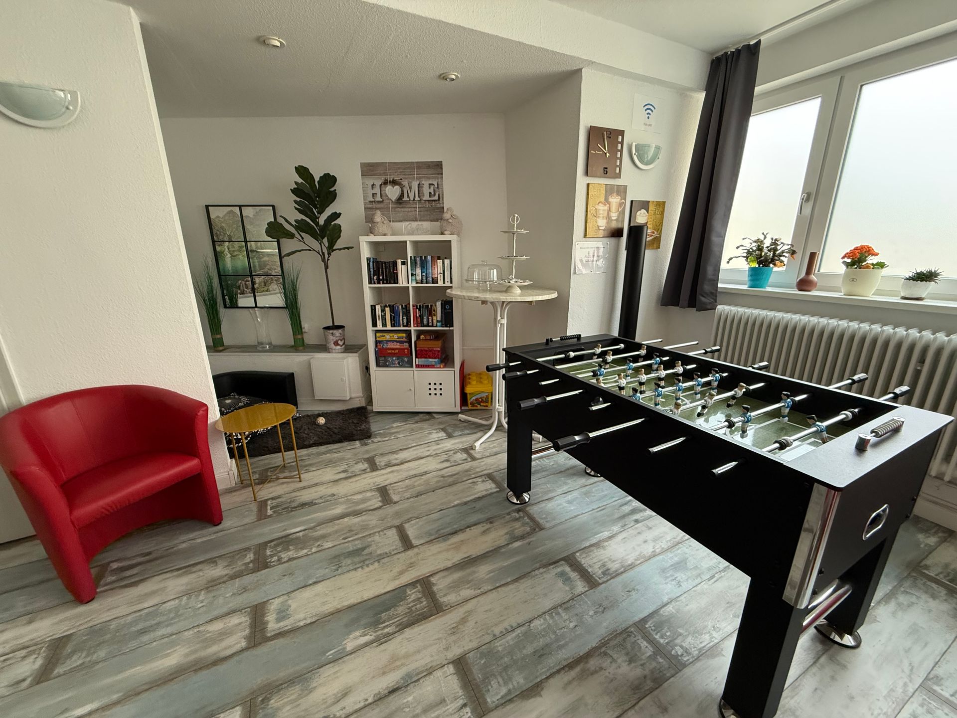 Wohnzimmer mit schwarzem Tischfußballtisch, rotem Sessel, Regalen und einem hellen Fenster zum Balkon.