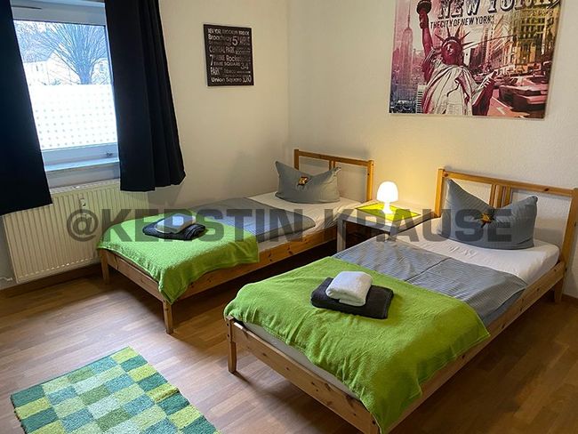 Zwei Einzelbetten mit grüner Bettwäsche in einem hellen Schlafzimmer, mit Nachttischlampe und Wandbildern.