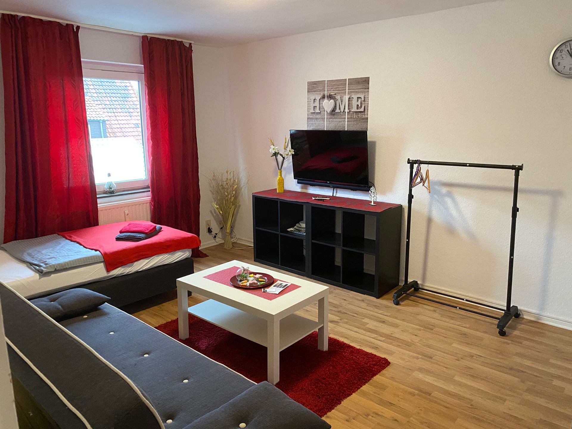 Kleines Wohnzimmer mit grauem Sofa, roten Vorhängen, Fernsehtisch, Couchtisch und Kleiderständer auf Holzboden