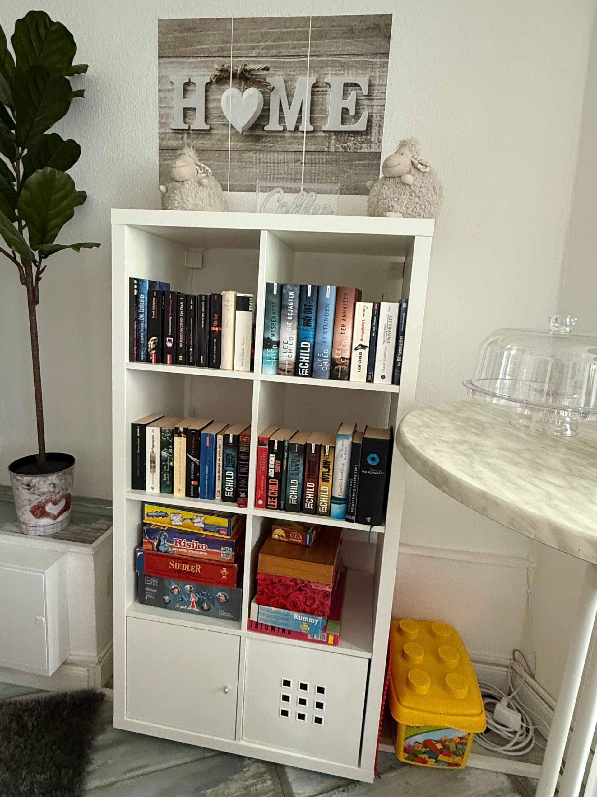 Weißes Bücherregal mit Büchern, Brettspielen und Körben neben einer kleinen Pflanze und einem runden Beistelltisch in einem hellen Raum