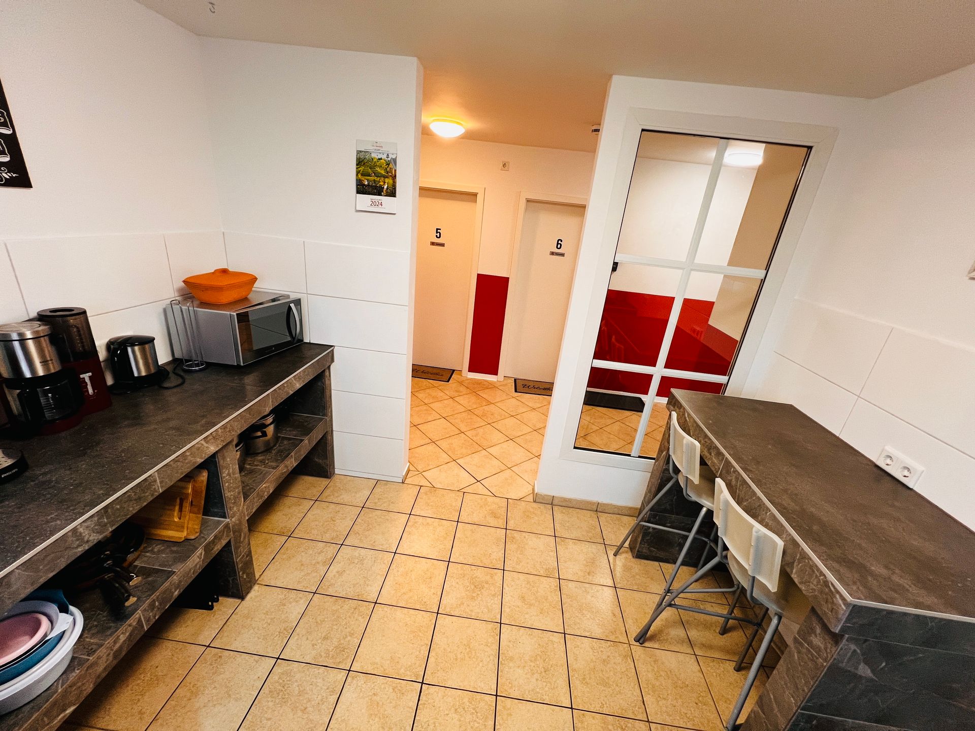 Kleine Küche mit Fliesenboden, Arbeitsflächen auf beiden Seiten und einer Tür, die zu einem rot-weißen Zimmer führt.