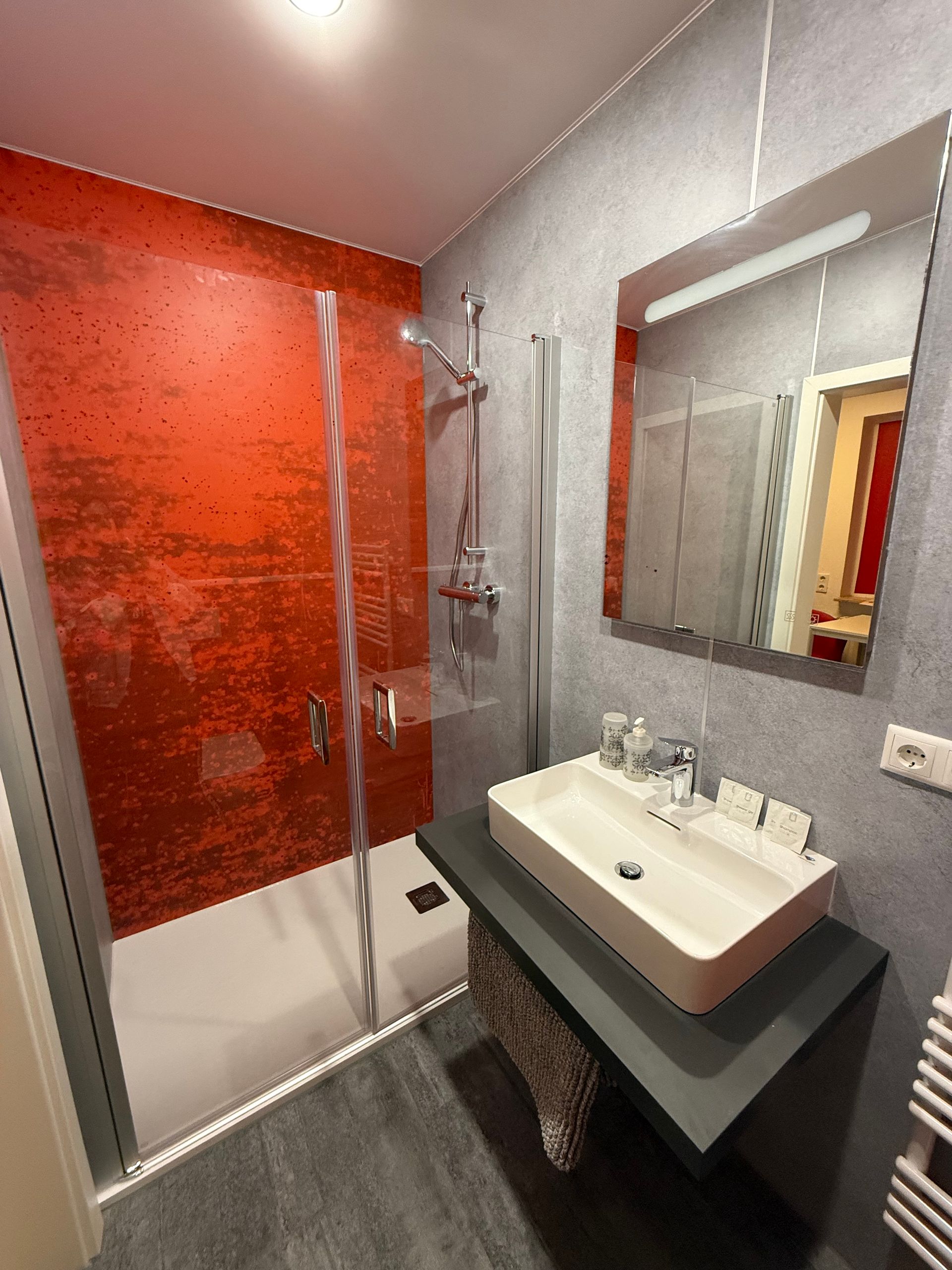 Modernes Badezimmer mit rot gefliester Dusche, grauen Wänden, Spiegel, Waschbecken und Handtuchheizkörper