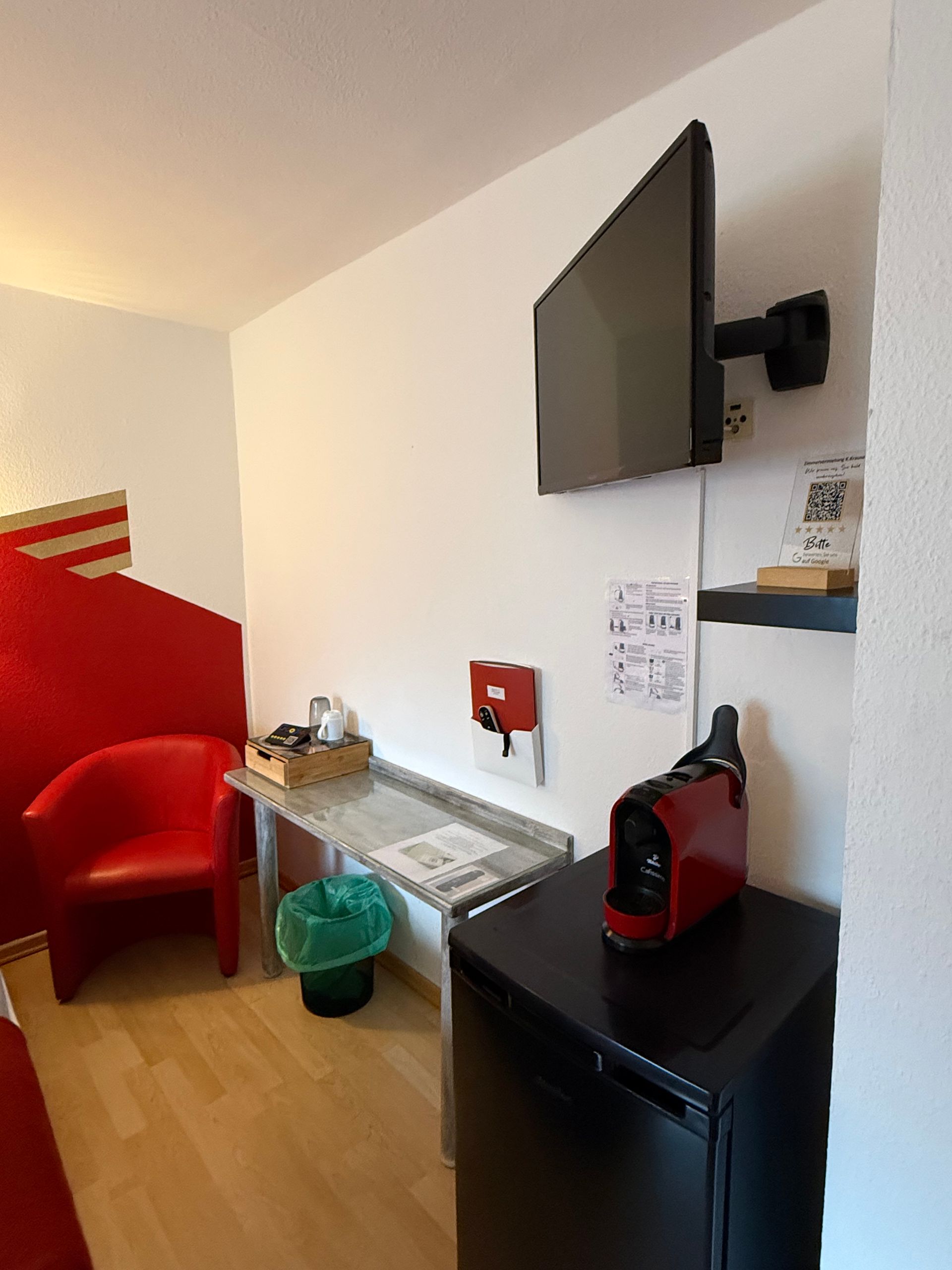 Kleines Hotelzimmer mit an der Wand montiertem Fernseher, Minikühlschrank, Schreibtisch, rotem Stuhl und grünem Mülleimer.