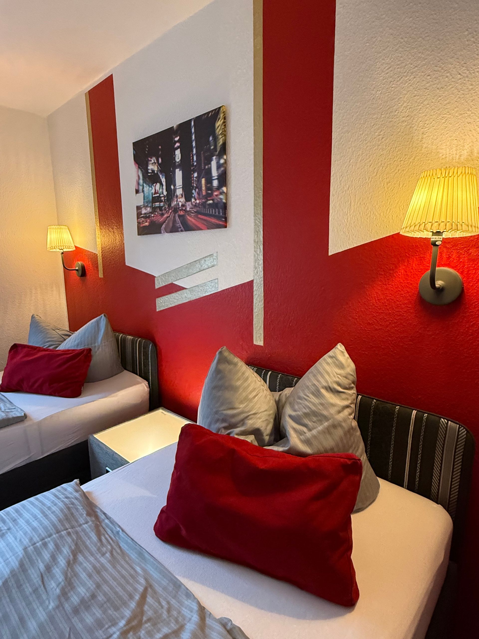 Gemütliches rot-weißes Schlafzimmer mit zwei Betten, Wandlampen und einem Stadtbilddruck