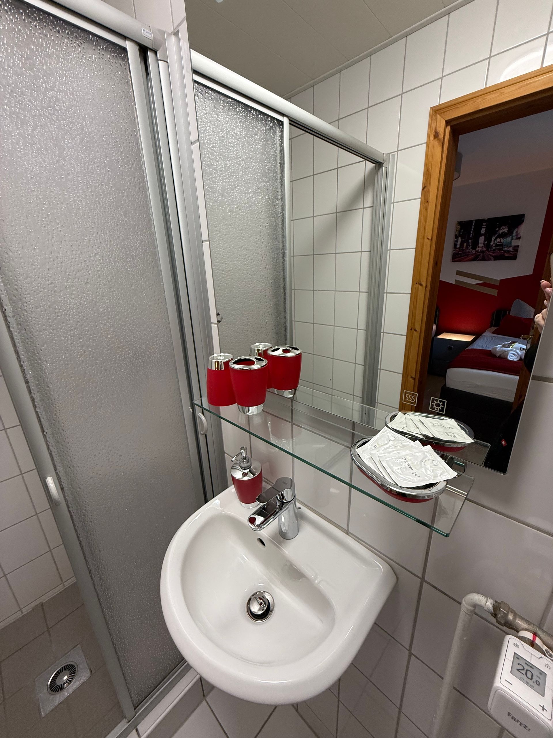 Kleines gefliestes Badezimmer mit Waschbecken, Duschvorhang, Spiegel und roten Seifenspendern über dem Waschbecken.