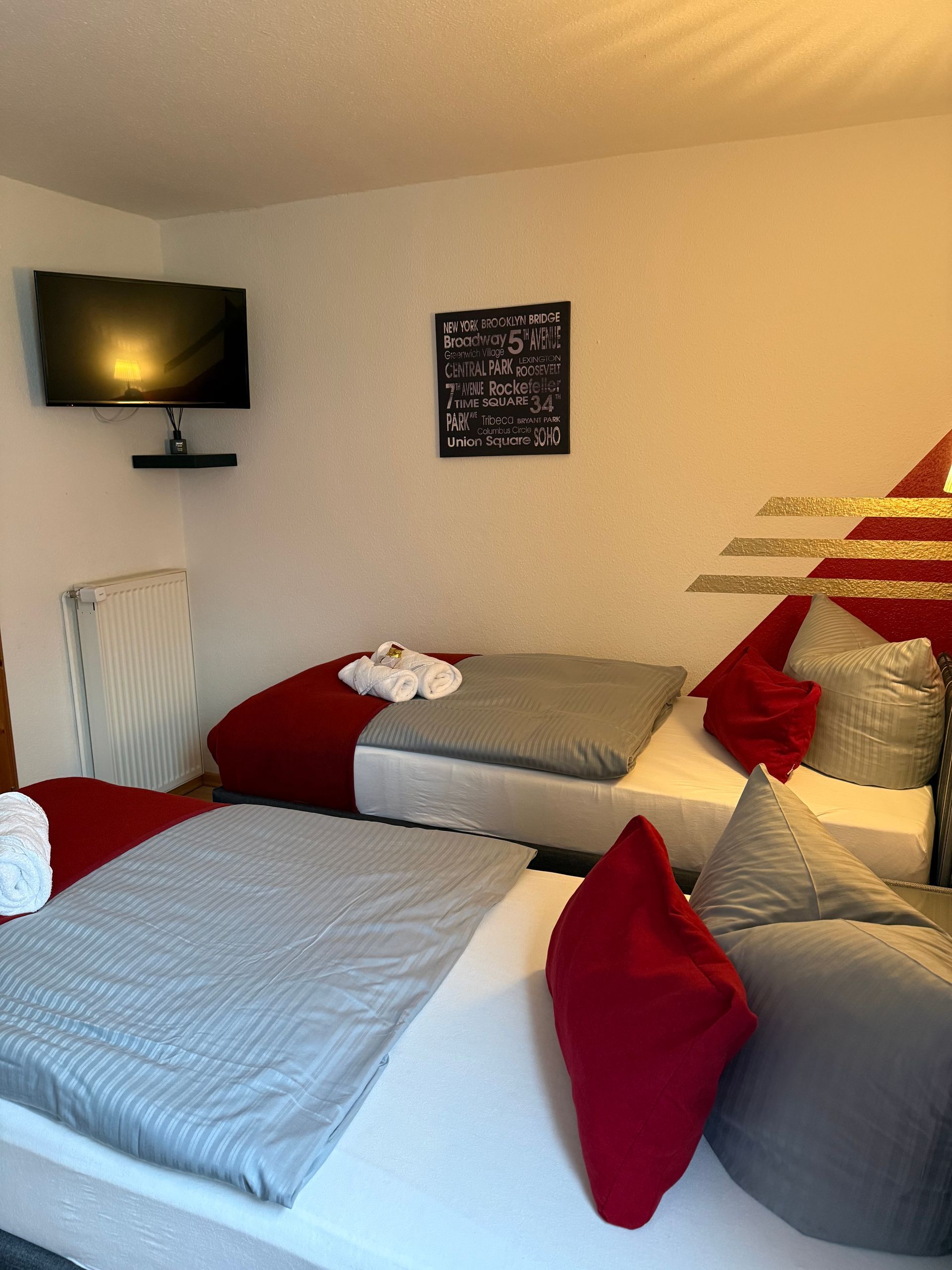 Kleines, gemütliches Schlafzimmer mit zwei Betten, roter und grauer Bettwäsche, an der Wand montiertem Fernseher und gerahmten Wandbildern.
