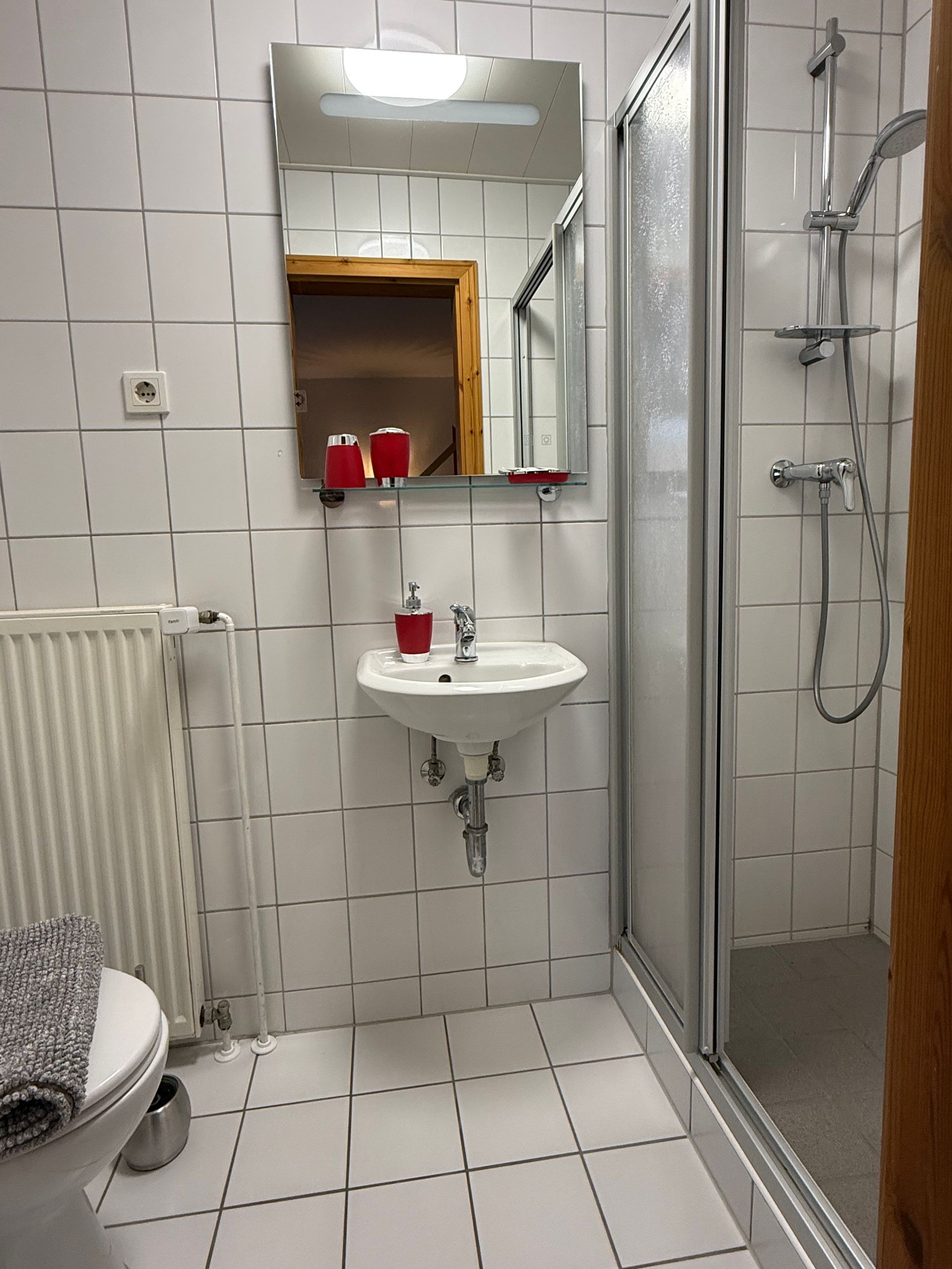 Kleines gefliestes Badezimmer mit Waschbecken, Spiegel, Duschkabine, Toilette und Heizkörper.