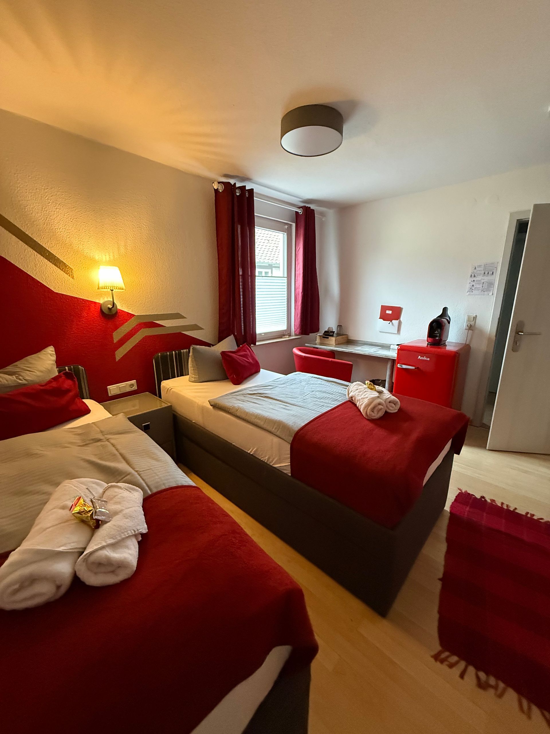 Rot-weißes Hotelzimmer mit zwei Betten, Nachttischlampe, Fenster und Kommode.