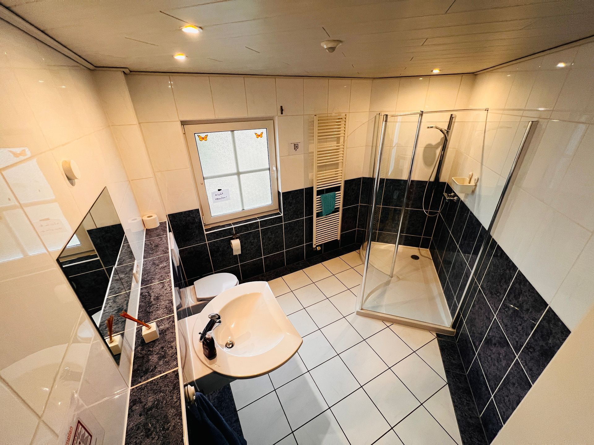 Kleines Badezimmer mit weißem Fliesenboden, schwarzen Wandfliesen, Waschbecken, Duschabtrennung und Fenster.