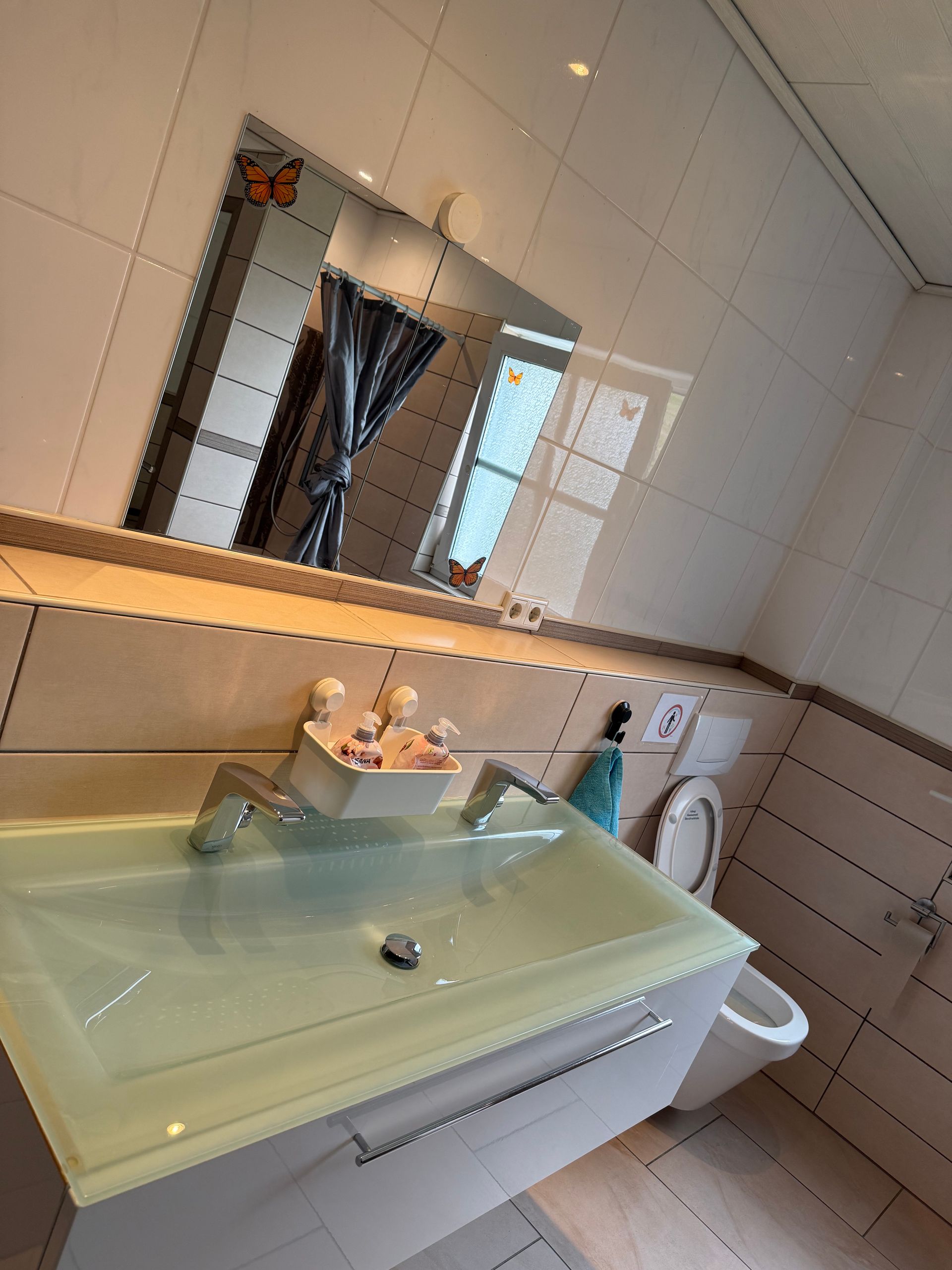 Badezimmer mit Glaswaschbecken, Spiegel, gefliesten Wänden und Toilette neben einem Fenster.