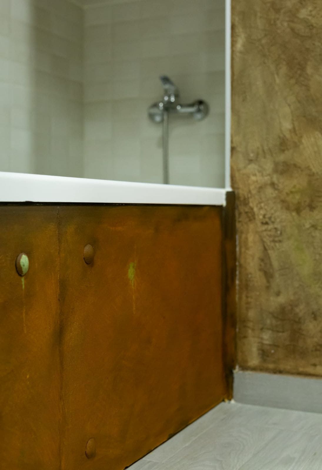 Baño con borde de bañera texturizado color óxido y pared dorada. Se ven el borde de la bañera y el grifo blancos.