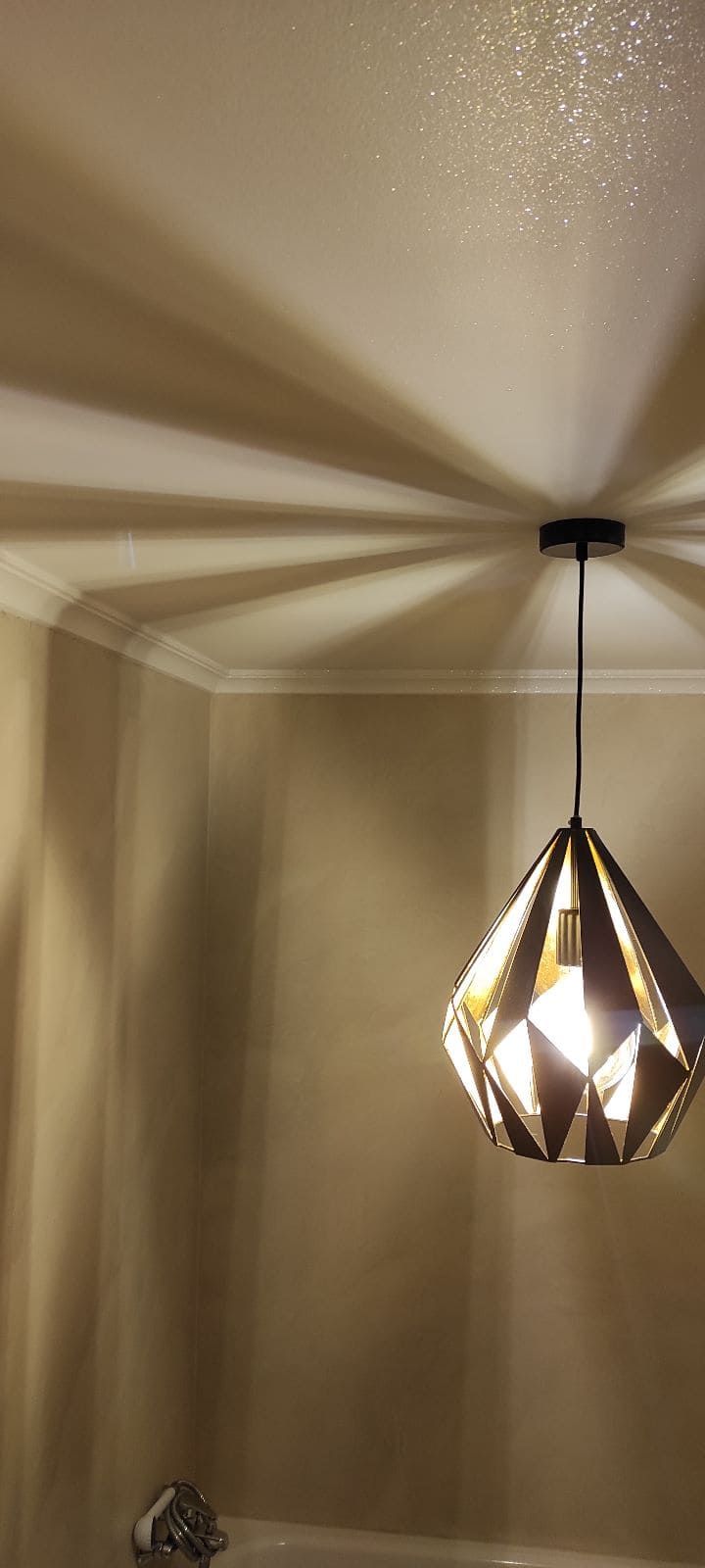 Una lámpara colgante en forma de diamante proyecta sombras geométricas en el techo y la pared de una habitación.