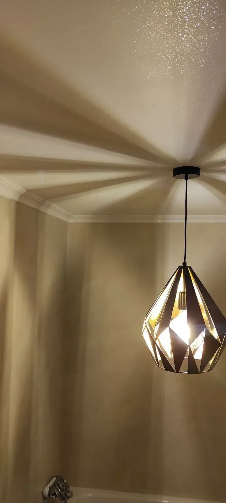 Lámpara colgante geométrica que proyecta sombras en forma de estrella sobre una pared y un techo de color beige.