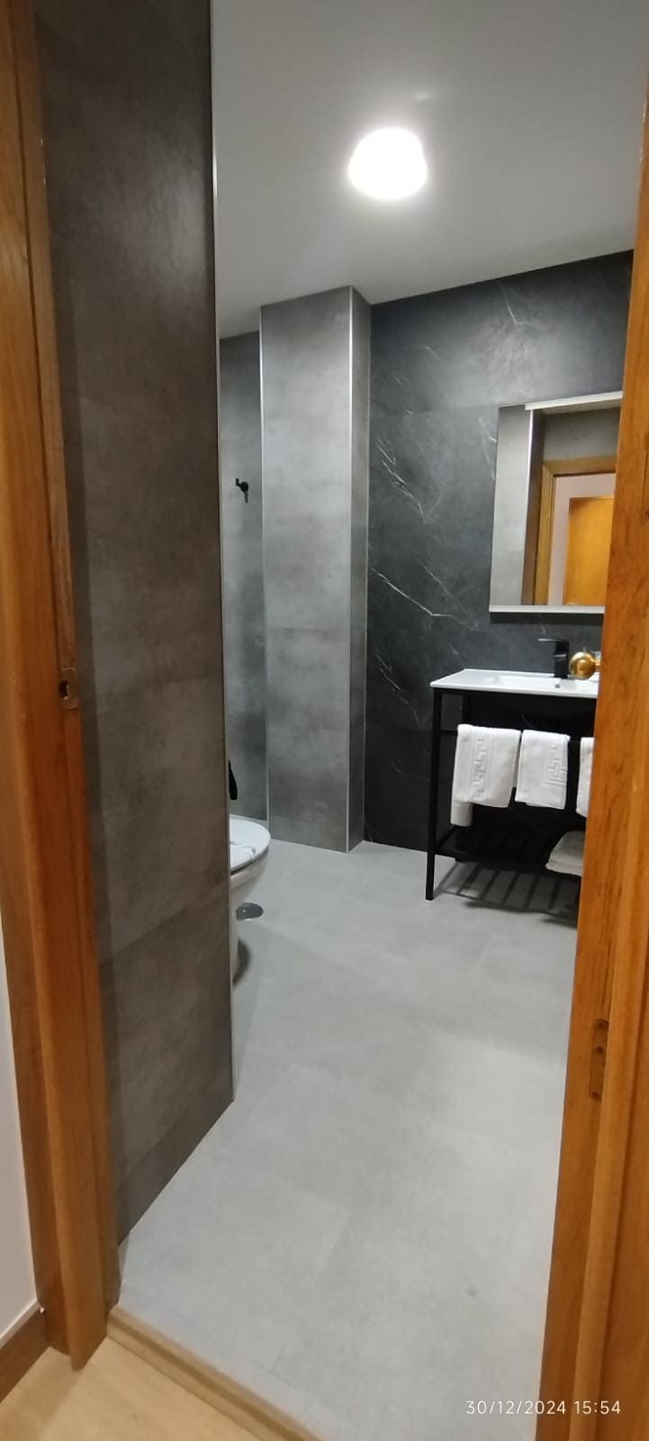 Interior de baño con paredes gris oscuro, piso gris, inodoro blanco y tocador negro con toallas blancas.