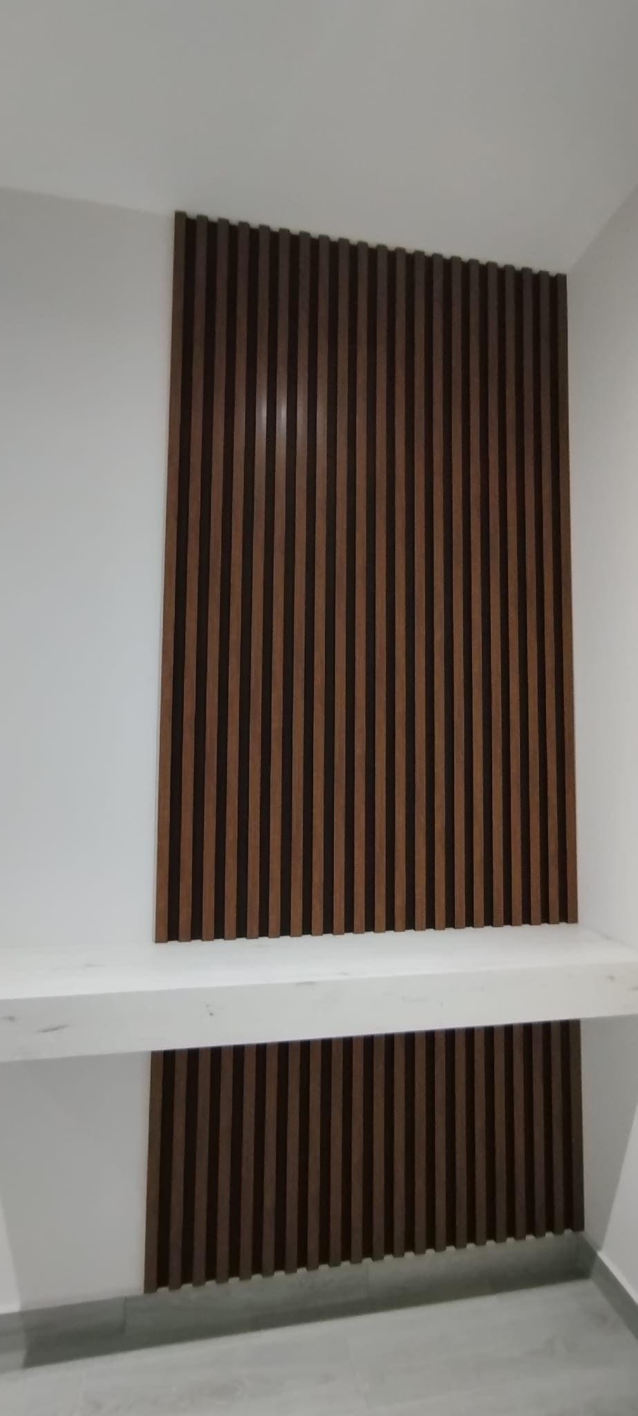 Pared de paneles de madera de color marrón con un estante blanco en una habitación con paredes blancas y suelo gris.
