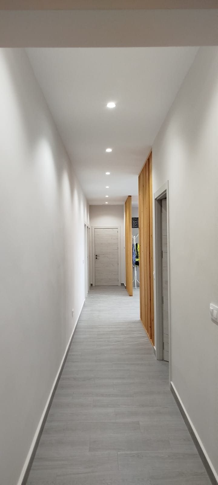 Un pasillo largo y bien iluminado con paredes blancas y suelo gris. Hay puertas y paneles de madera.
