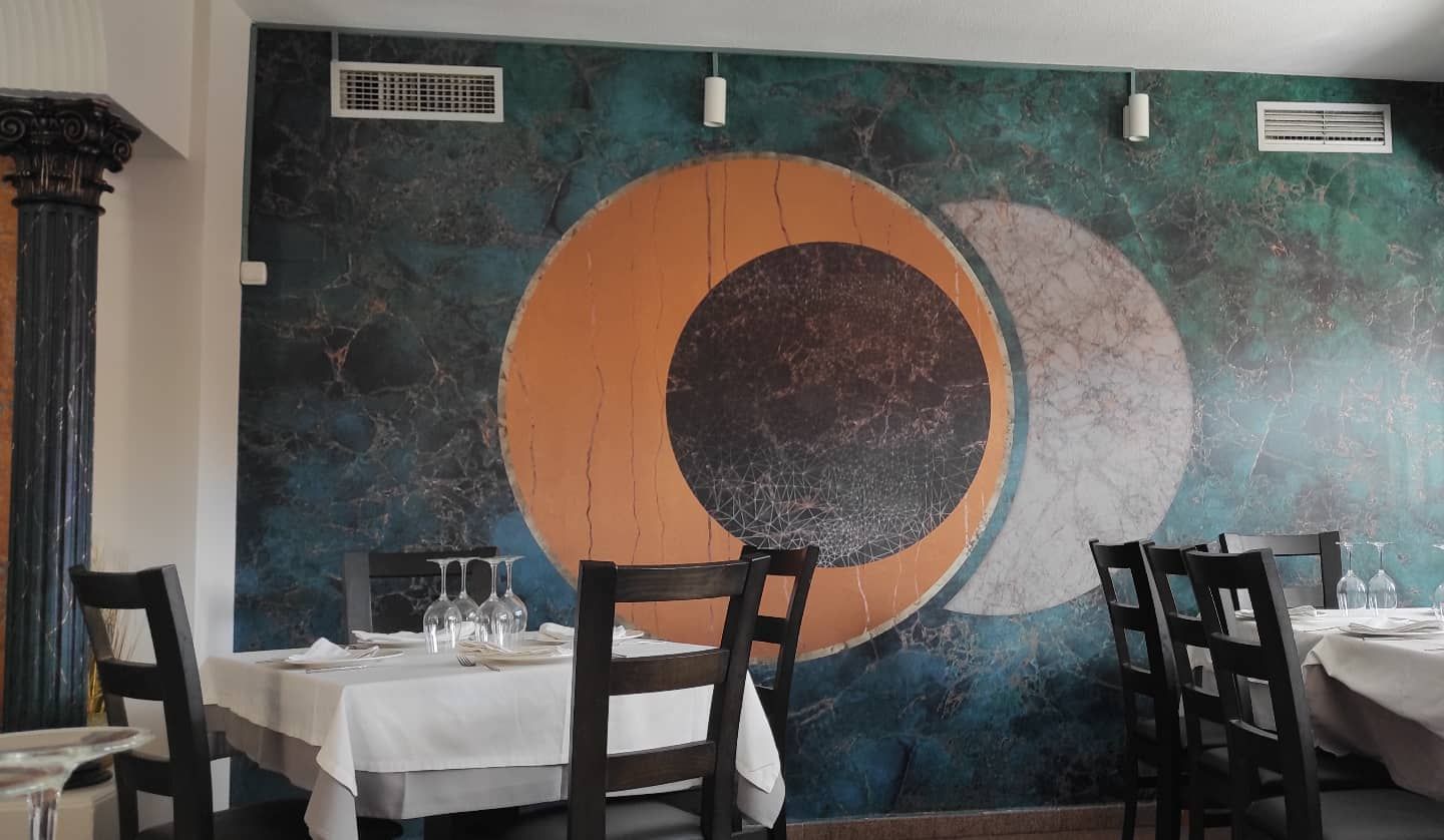 Interior de restaurante con un mural decorativo de círculos superpuestos en naranja y gris, mesas preparadas para cenar.