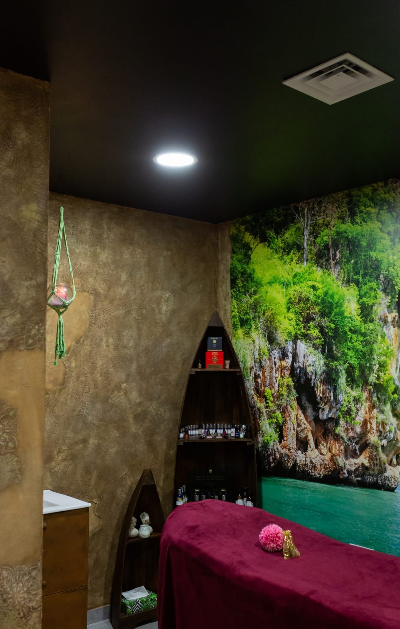 Pequeña sala de tratamientos de spa con camilla de masaje, estantes y un mural tropical en la pared.
