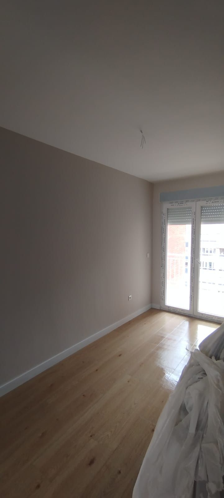 Habitación vacía con suelo de madera clara, paredes de color beige y puertas de cristal que dan a un balcón.