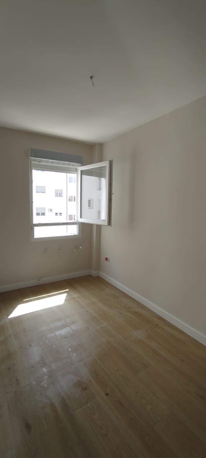 Habitación blanca vacía con suelo de madera, ventana abierta y luz solar que entra a raudales por el suelo.