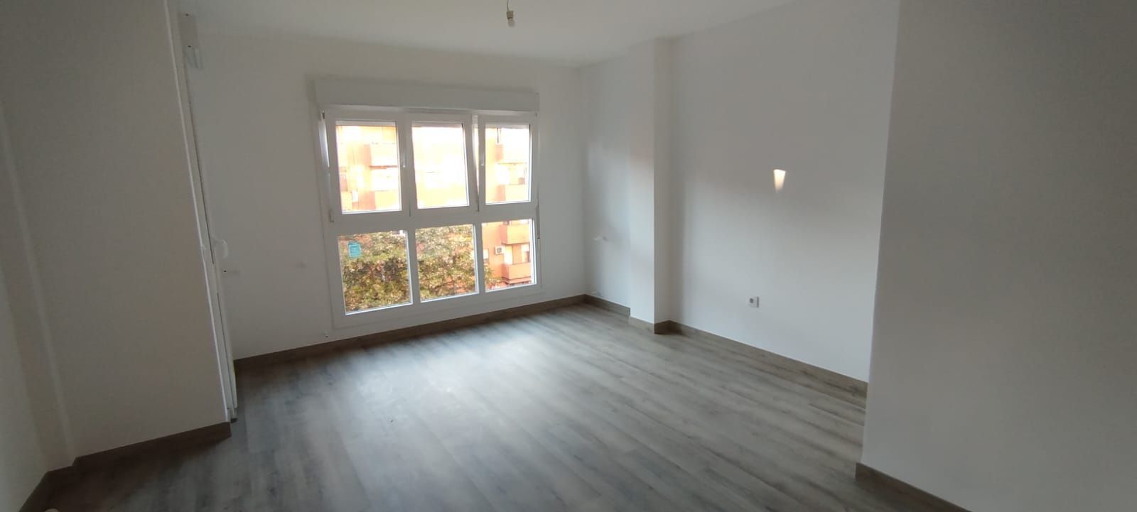 Habitación blanca vacía con suelo gris y una gran ventana que deja entrar la luz del día.