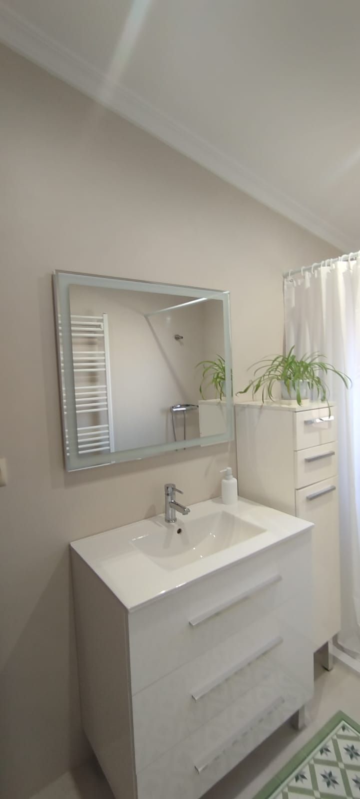 Tocador de baño luminoso con espejo, lavabo y armarios blancos junto a una ventana con cortinas transparentes.