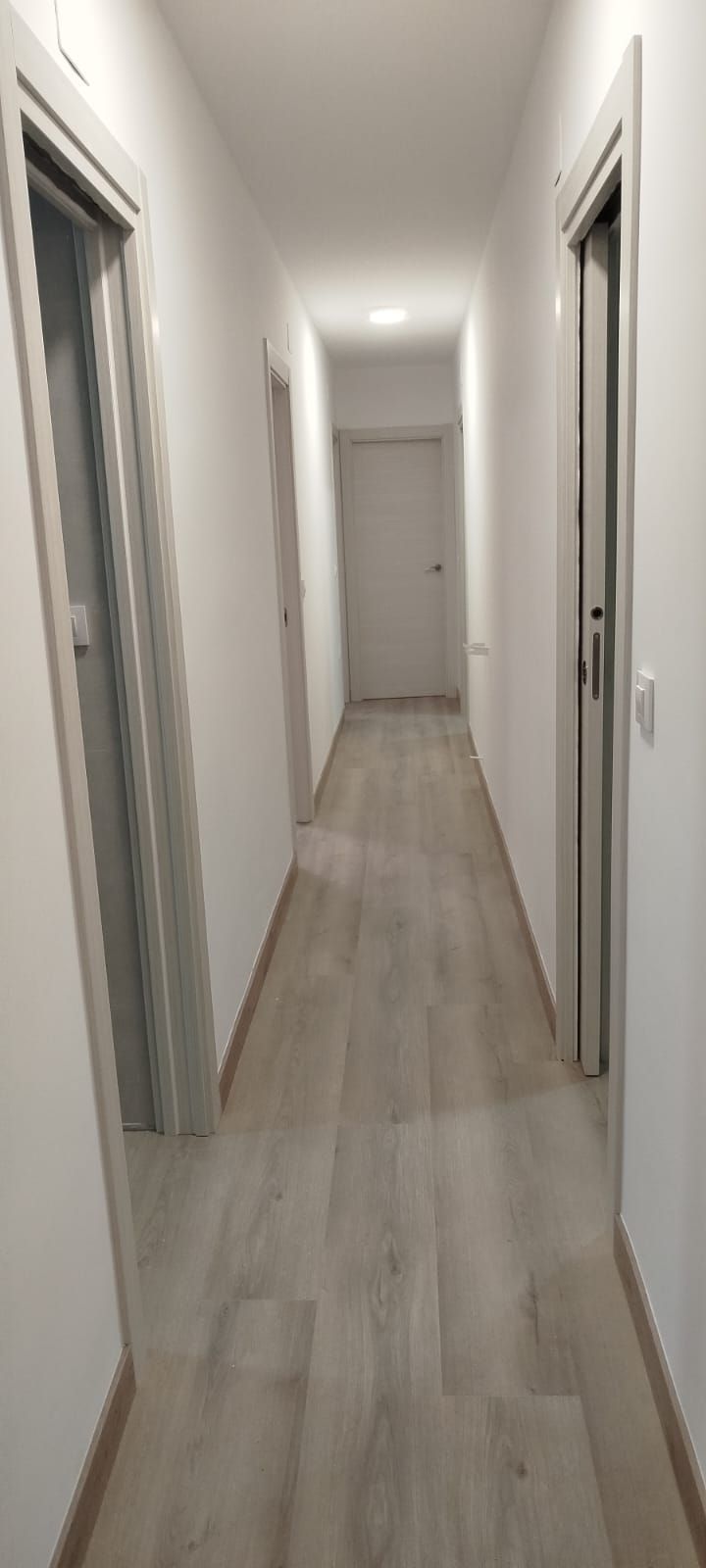 Pasillo largo y estrecho con paredes blancas, suelos de madera clara y puertas a ambos lados.
