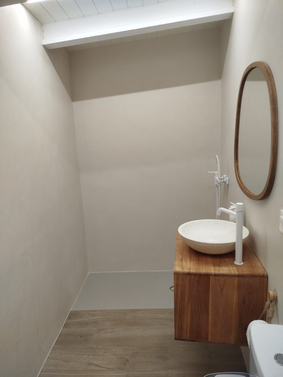 Baño pequeño y minimalista con techo inclinado, tocador de madera, espejo redondo y lavabo blanco sobre encimera.