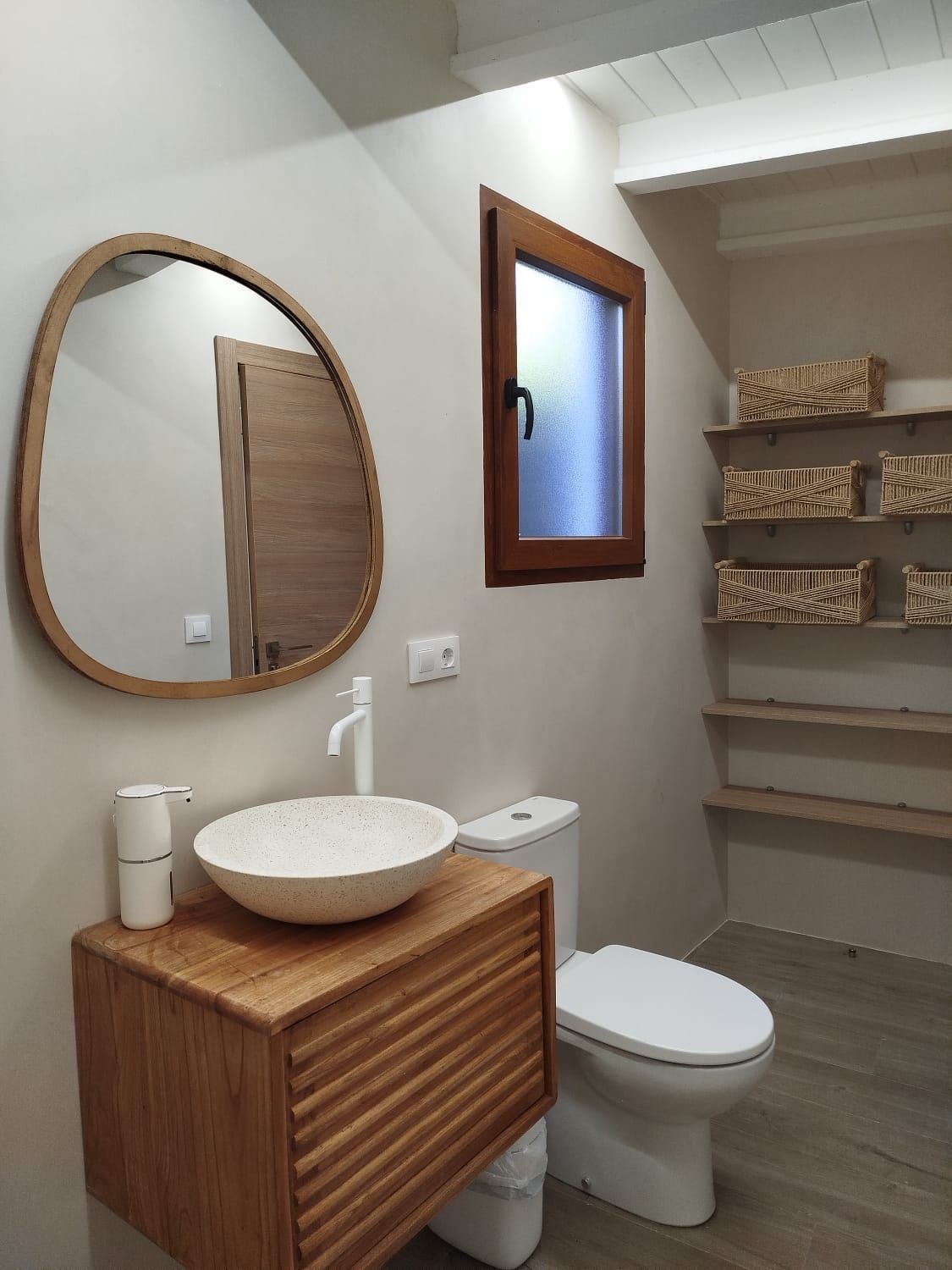 Baño moderno con tocador de madera, espejo redondo, inodoro y estantes abiertos debajo de una ventana.