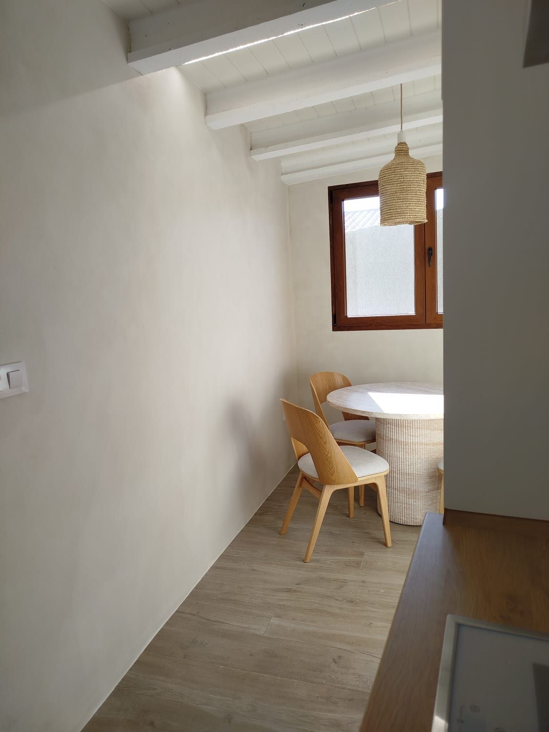 Un estrecho rincón para comer con sillas de madera, mesa redonda, ventana y lámpara colgante en una habitación blanca.