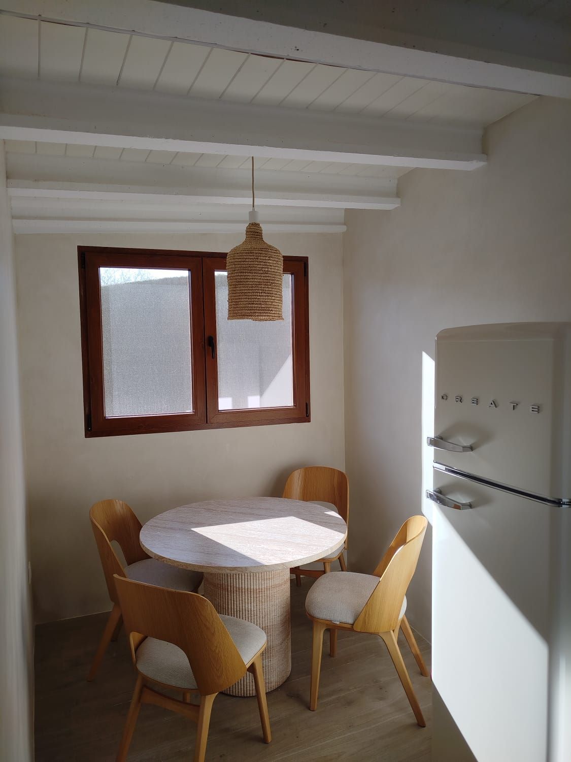 Pequeño rincón comedor con mesa redonda blanca, cuatro sillas de madera, ventana y lámpara colgante tejida.