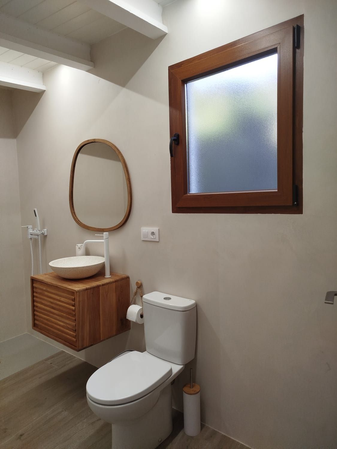Baño pequeño con inodoro, lavabo de madera, espejo ovalado y ventana esmerilada.