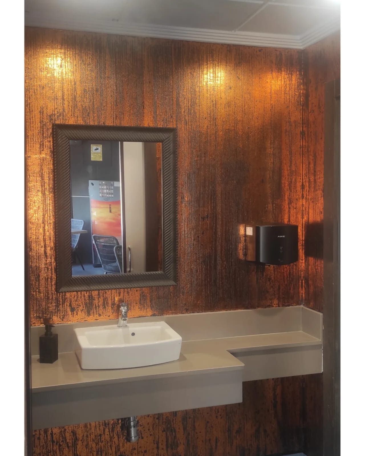 Baño con pared texturizada color cobre, espejo rectangular, lavabo blanco y dispensador de jabón negro.