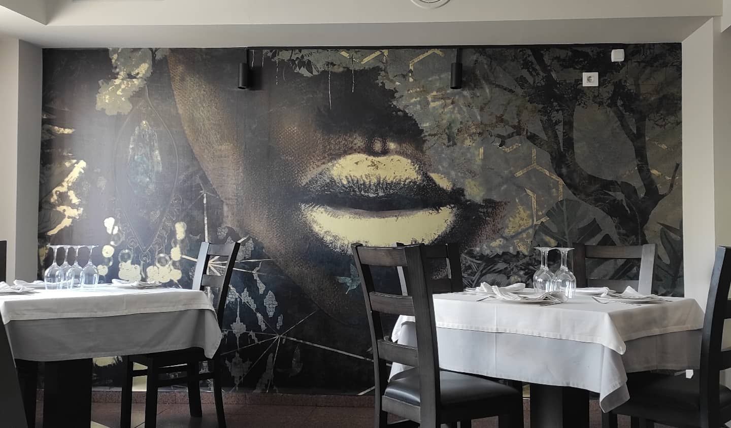 Interior de restaurante con mesas preparadas para cenar, sillas oscuras y un mural de una cara en la pared.