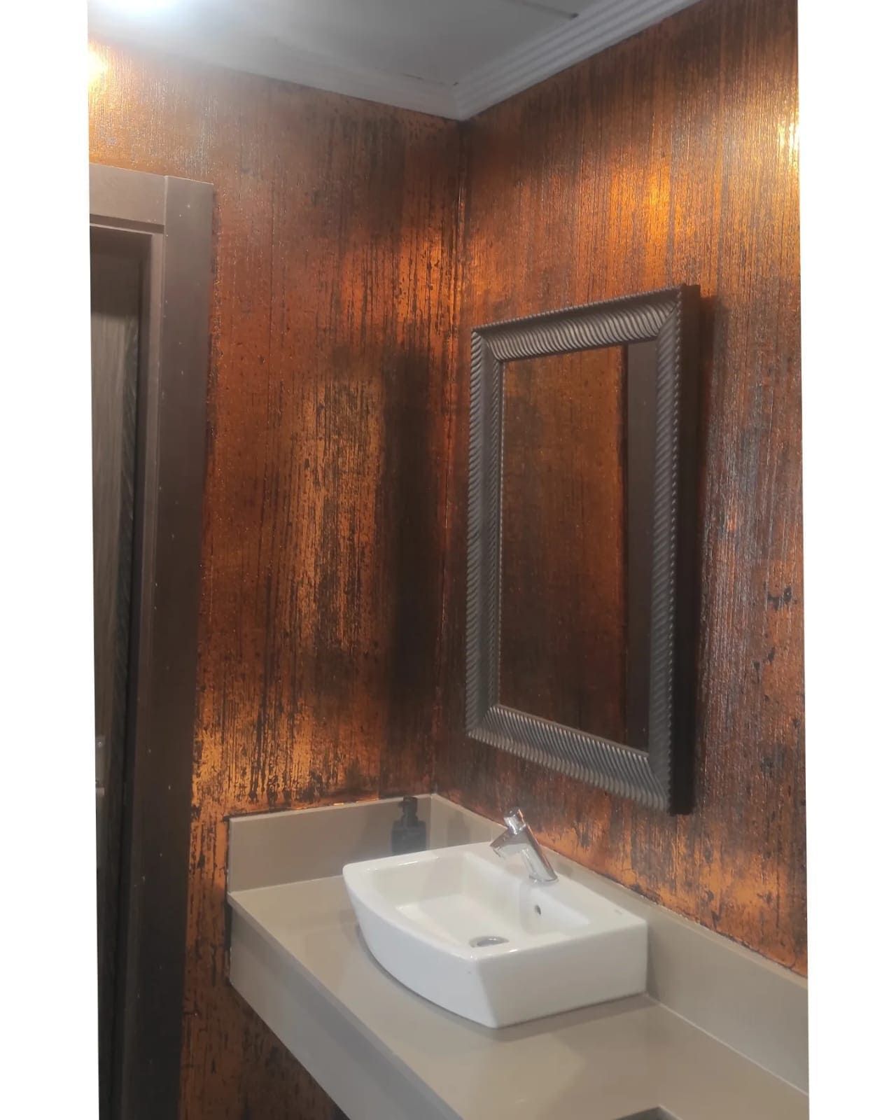 Baño con paredes texturizadas color cobre, lavabo blanco y espejo enmarcado.