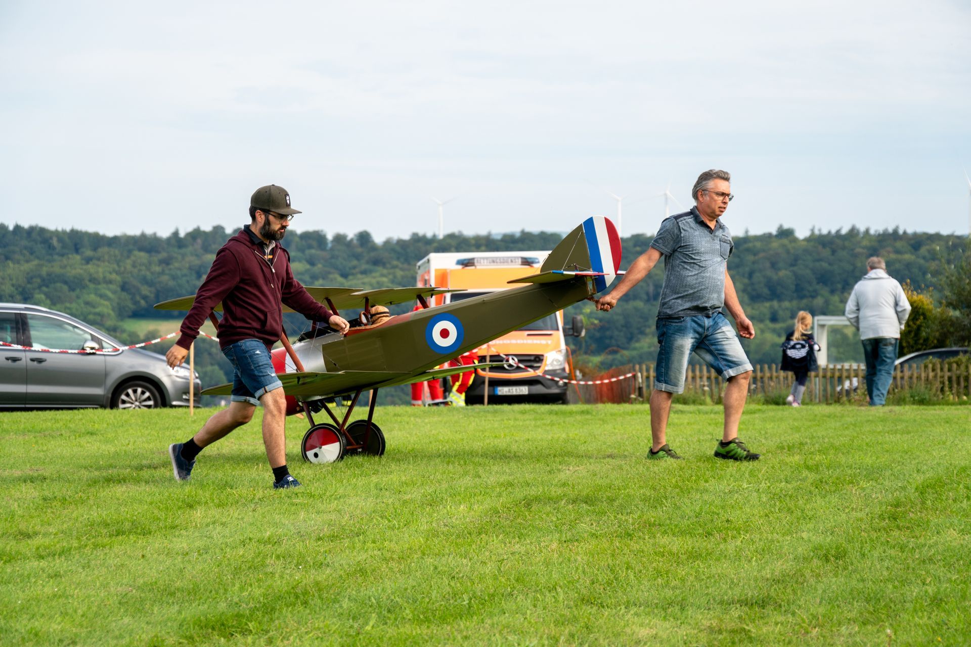 Zwei Männer tragen ein Modellflugzeug über ein grasbewachsenes Feld. Das Flugzeug ist olivgrün mit einer blau-weißen Kokarde.
