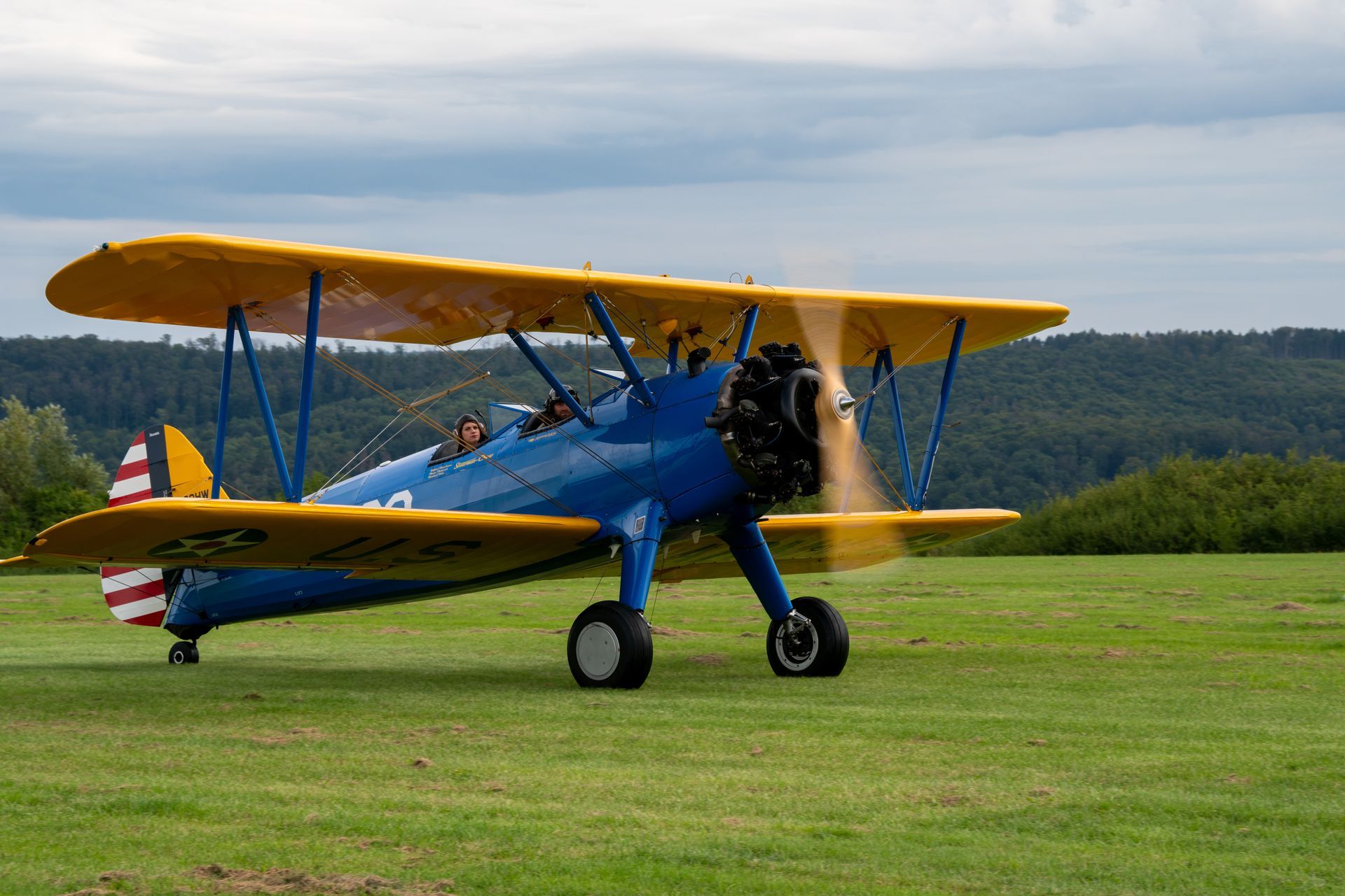 Blau-gelbes Doppeldeckerflugzeug auf einem Grasfeld, Propeller dreht sich. US-Flagge am Heck.