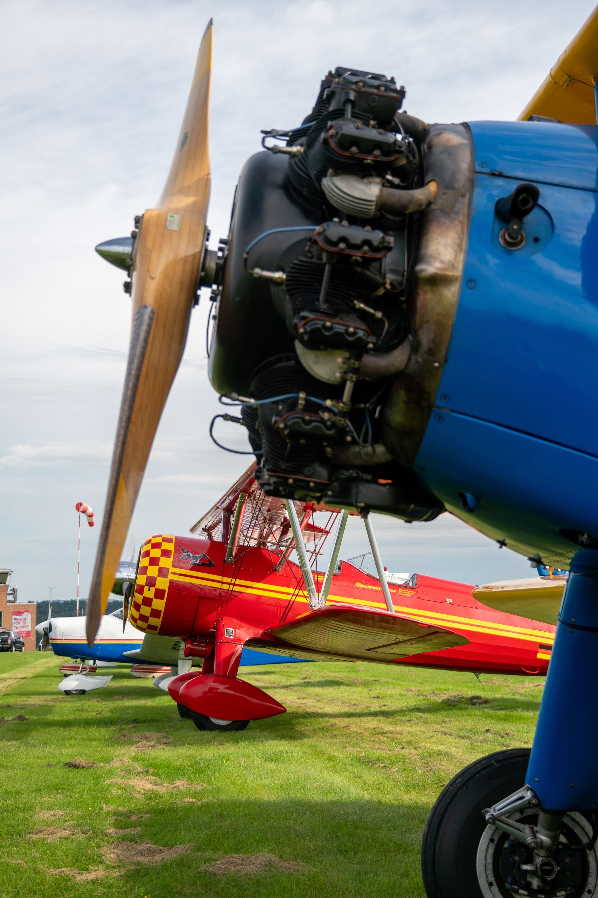 Nahaufnahme eines blauen Doppeldeckermotors mit Holzpropeller, im Hintergrund ein rot-gelber Doppeldecker auf einem grasbewachsenen Feld.