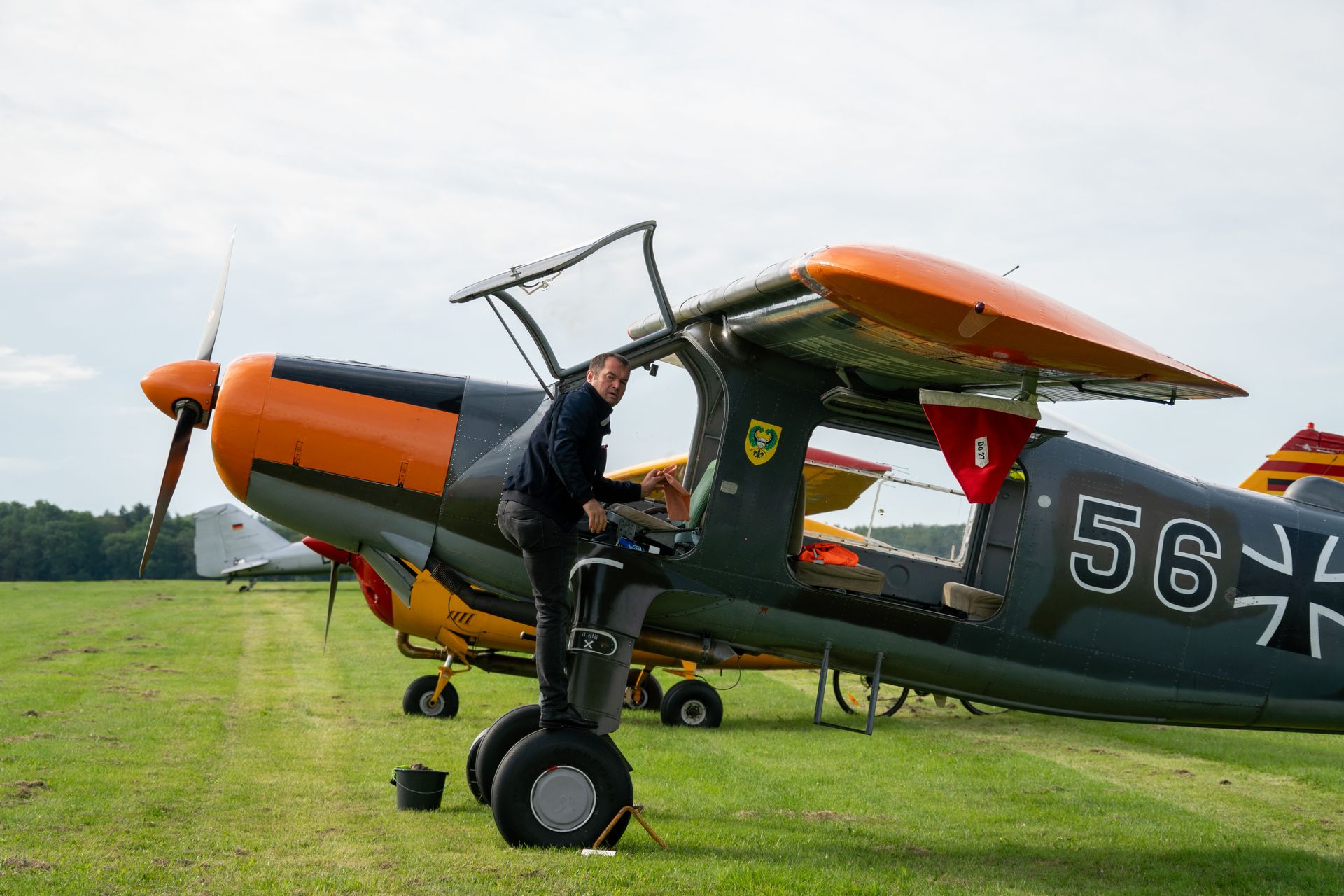 Mann neben einem orange-grauen Doppeldecker mit schwarzen Rädern auf einem grasbewachsenen Feld.