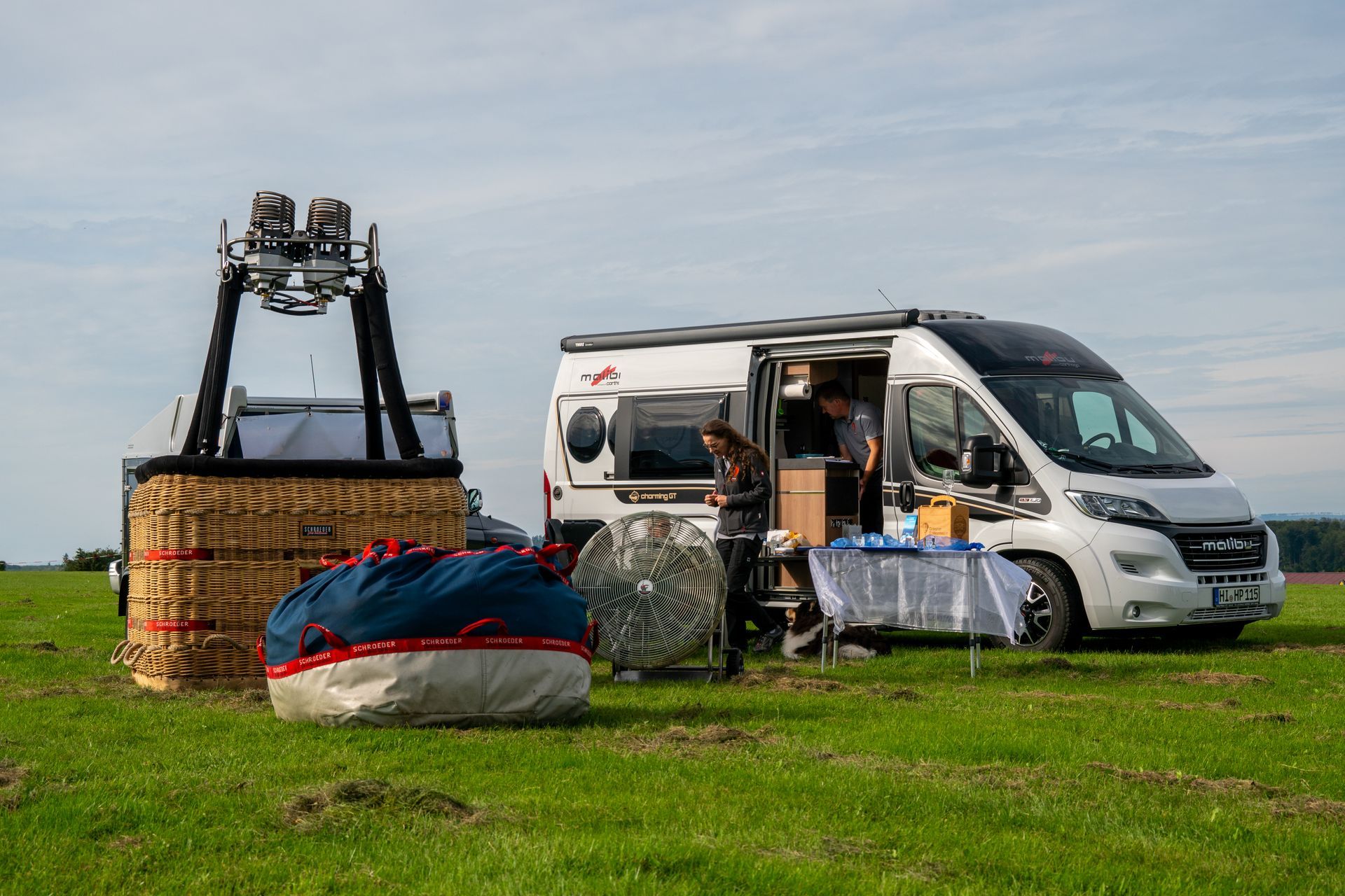 Auf einer Wiese steht ein Heißluftballon mit einem Wohnmobil. Die Leute bereiten sich auf die Ballonfahrt vor.