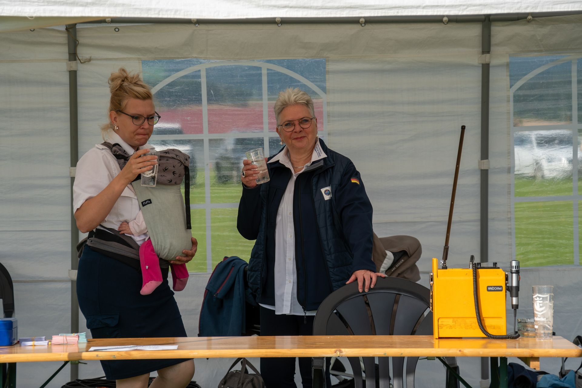 Zwei Frauen sitzen an einem Tisch unter einem Baldachin. Die eine trägt eine Babytrage und hält ein Getränk. Die andere trinkt etwas.
