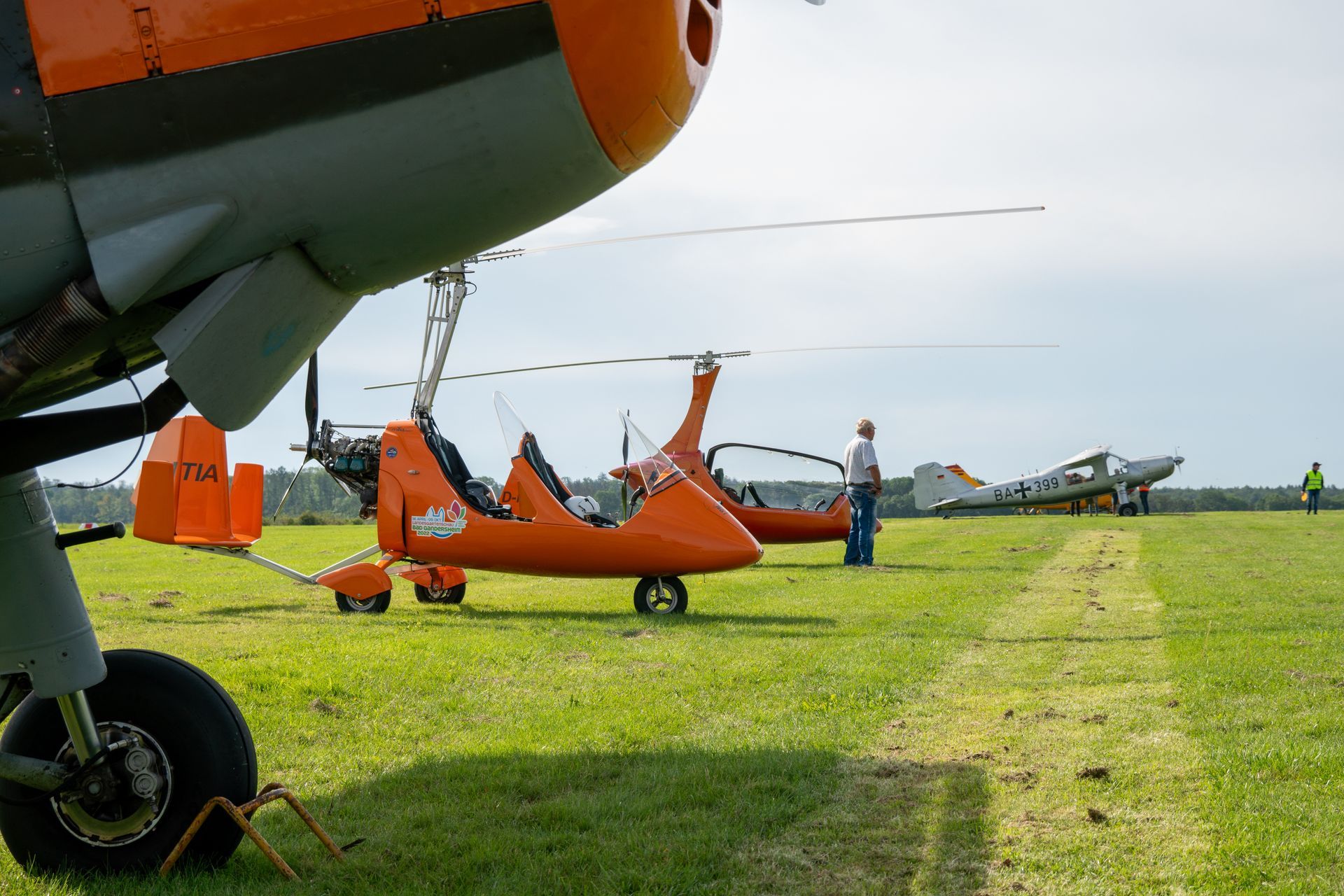 Ein orangefarbenes Flugzeug und ein Tragschrauber sind auf einem grasbewachsenen Flugfeld geparkt, eine Person steht in der Nähe.